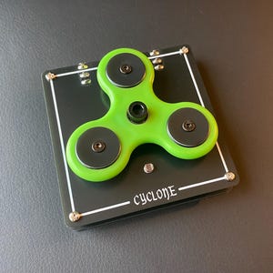 May include: A green and black fidget spinner with the word "CYCLONE" printed on the base. The spinner has three arms with black circular weights and is mounted on a black square base with white trim and silver screws.