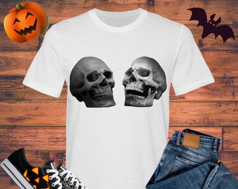 May include: White t-shirt with a graphic of two black and white skulls facing each other. The skulls are detailed and have a realistic look. The t-shirt is on a wooden background with a pair of black and orange sneakers and blue jeans.