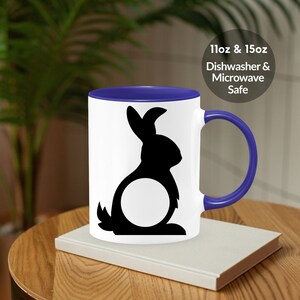 May include: A white ceramic mug with a blue rim and handle. The mug features a black silhouette of a rabbit with a circle in the middle. The text "11oz & 15oz Dishwasher & Microwave Safe" is printed on the mug.