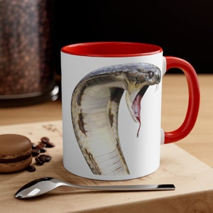 Könnte beinhalten: Weiße Keramik-Kaffeetasse mit rotem Rand und Henkel. Die Tasse zeigt ein Motiv einer Kobra mit offenem Maul, die ihre Zähne zeigt. Die Schlange ist braun und weiß mit einem strukturierten Muster.