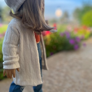 Op de afbeelding: Een pop die een witte gebreide cardigan, blauwe jeans en een witte gebreide muts draagt. De pop staat op een stoep met een wazige achtergrond van bloemen.