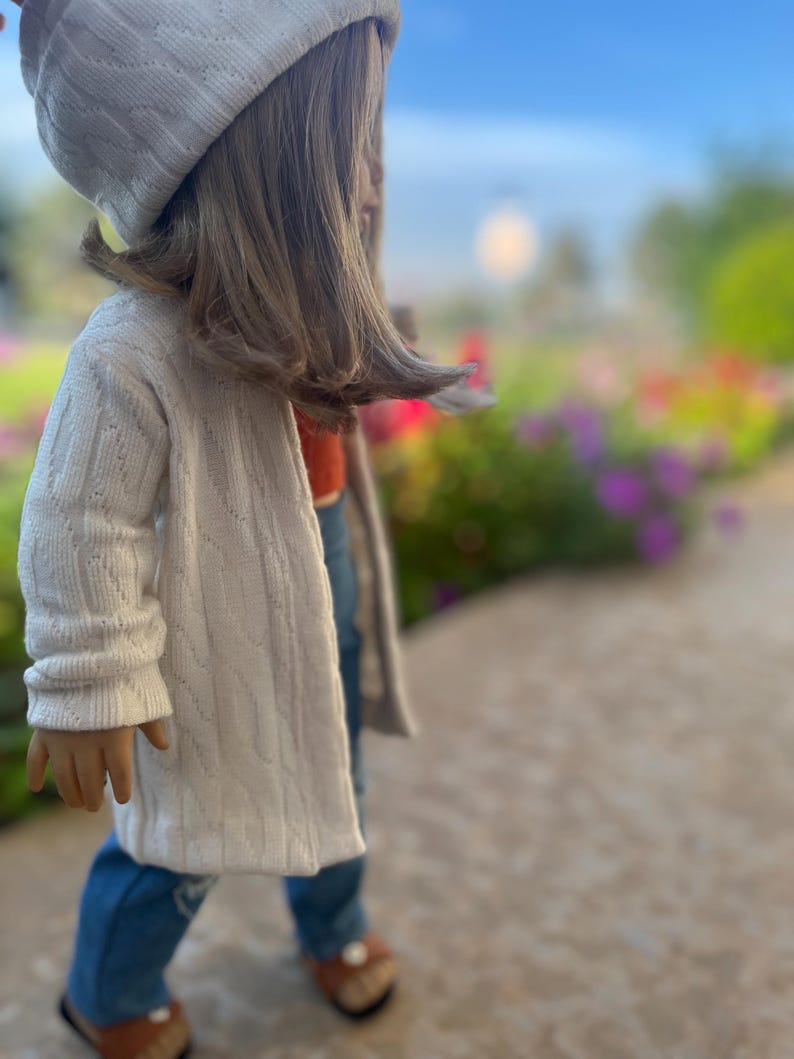 Op de afbeelding: Een pop die een witte gebreide cardigan, blauwe jeans en een witte gebreide muts draagt. De pop staat op een stoep met een wazige achtergrond van bloemen.