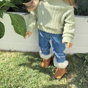 Puede incluir: Una mu&ntilde;eca con un su&eacute;ter verde claro, jeans azules con dobladillos deshilachados y botas de piel sint&eacute;tica marr&oacute;n.