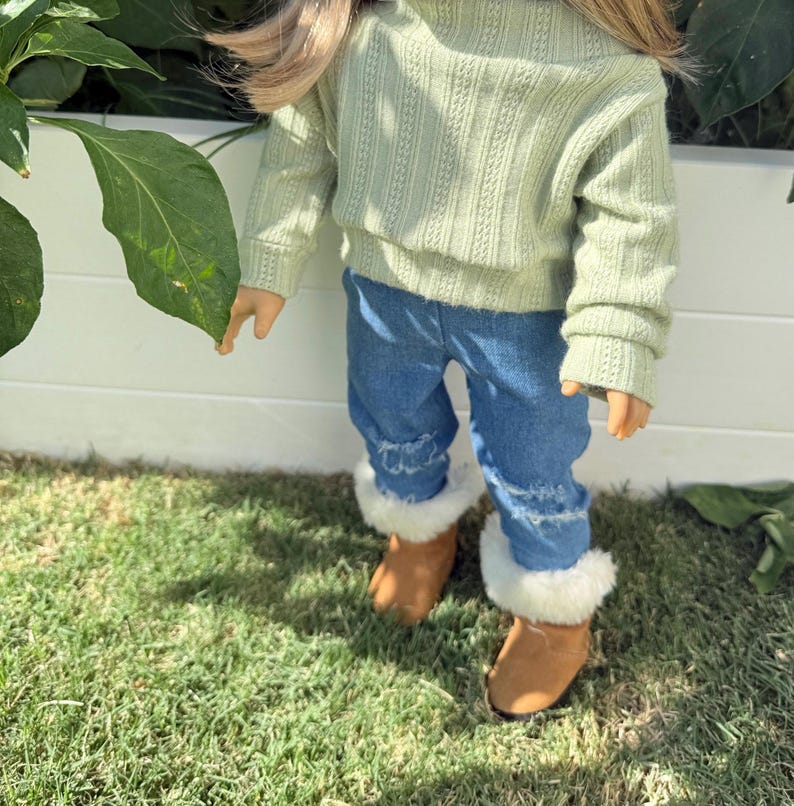 Puede incluir: Una mu&ntilde;eca con un su&eacute;ter verde claro, jeans azules con dobladillos deshilachados y botas de piel sint&eacute;tica marr&oacute;n.