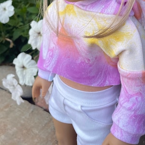 K&ouml;nnte beinhalten: Ein rosa, gelber und wei&szlig;er tie-dye-Cropped-Hoodie mit langen &Auml;rmeln. Der Hoodie wird &uuml;ber einer wei&szlig;en Shorts getragen.