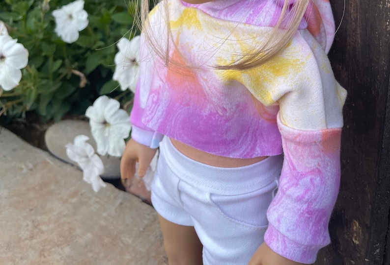 K&ouml;nnte beinhalten: Ein rosa, gelber und wei&szlig;er tie-dye-Cropped-Hoodie mit langen &Auml;rmeln. Der Hoodie wird &uuml;ber einer wei&szlig;en Shorts getragen.