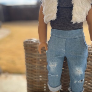 May include: A doll wearing a brown turtleneck shirt with a white faux fur vest, blue jeans with distressed details, and white shoes.