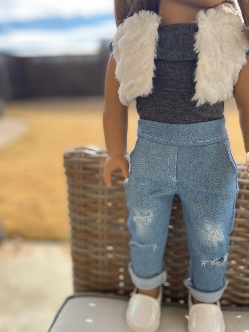 May include: A doll wearing a brown turtleneck shirt with a white faux fur vest, blue jeans with distressed details, and white shoes.