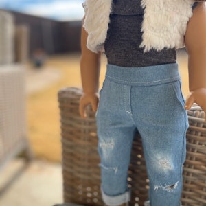 May include: A doll wearing a gray mock neck shirt, a white faux fur vest, blue jeans with distressed details, and white shoes.
