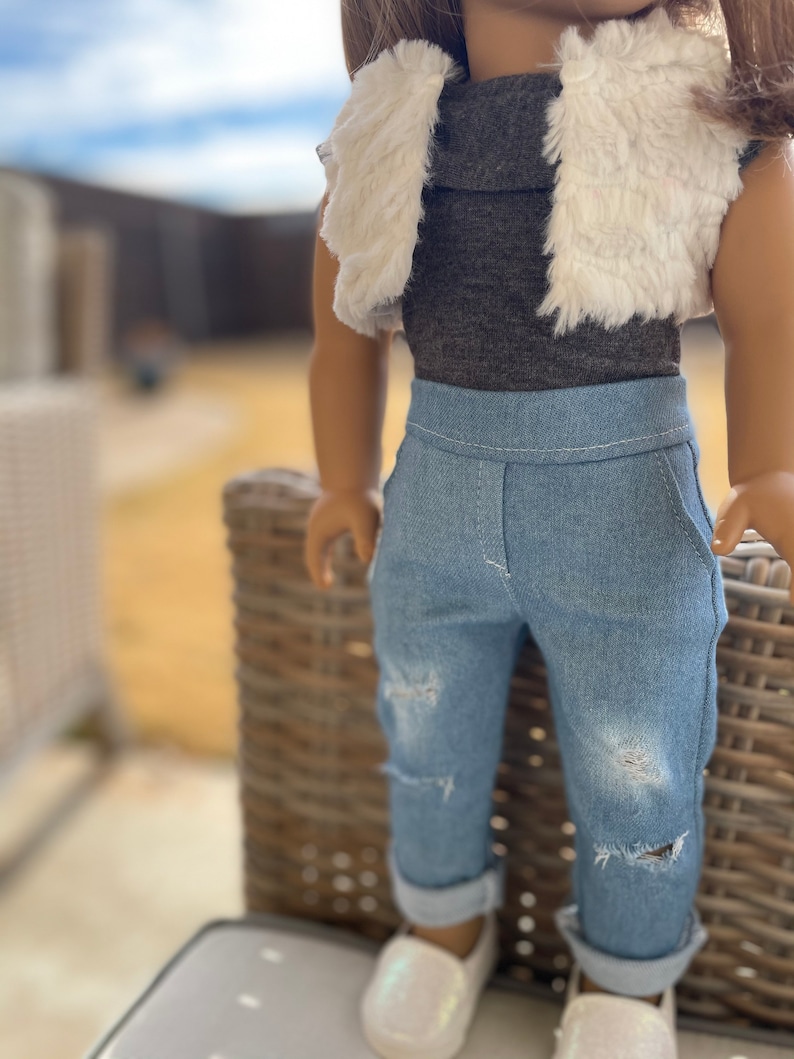 May include: A doll wearing a gray mock neck shirt, a white faux fur vest, blue jeans with distressed details, and white shoes.