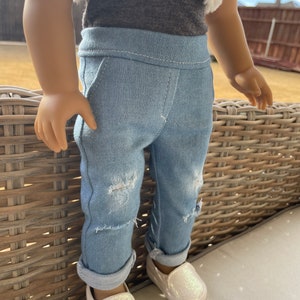 May include: A pair of light blue denim jeans with distressed details and rolled cuffs. The jeans are worn by a doll and are paired with white sparkly slip-on shoes.