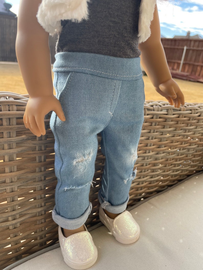 May include: A pair of light blue denim jeans with distressed details and rolled cuffs. The jeans are worn by a doll and are paired with white sparkly slip-on shoes.