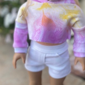 K&ouml;nnte beinhalten: Ein tie-dye-farbener, verk&uuml;rzter Hoodie mit langen &Auml;rmeln und einem wei&szlig;en Bund. Der Hoodie wird mit einer wei&szlig;en Shorts kombiniert.