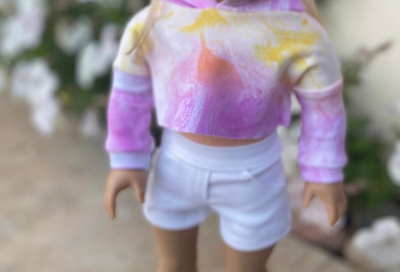 K&ouml;nnte beinhalten: Ein tie-dye-farbener, verk&uuml;rzter Hoodie mit langen &Auml;rmeln und einem wei&szlig;en Bund. Der Hoodie wird mit einer wei&szlig;en Shorts kombiniert.