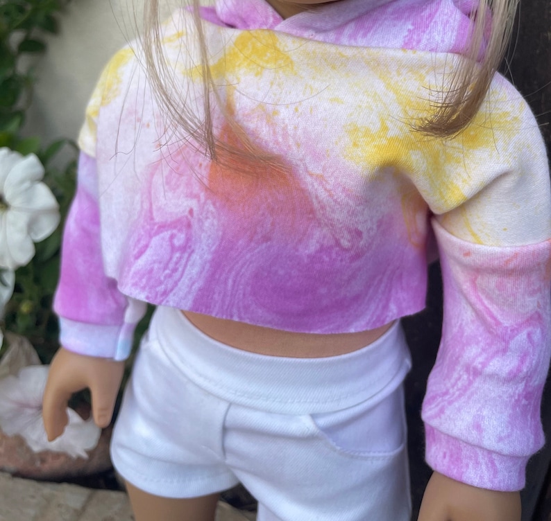 K&ouml;nnte beinhalten: Ein tie-dye-farbener, kurzer Hoodie mit langen &Auml;rmeln in den Farben Pink, Gelb und Wei&szlig;. Der Hoodie hat eine lockere Passform und einen l&auml;ssigen Stil.