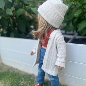 Op de afbeelding: Een pop die een witte kabelgebreide cardigan draagt, een rood topje, blauwe jeans en een witte kabelgebreide beanie.