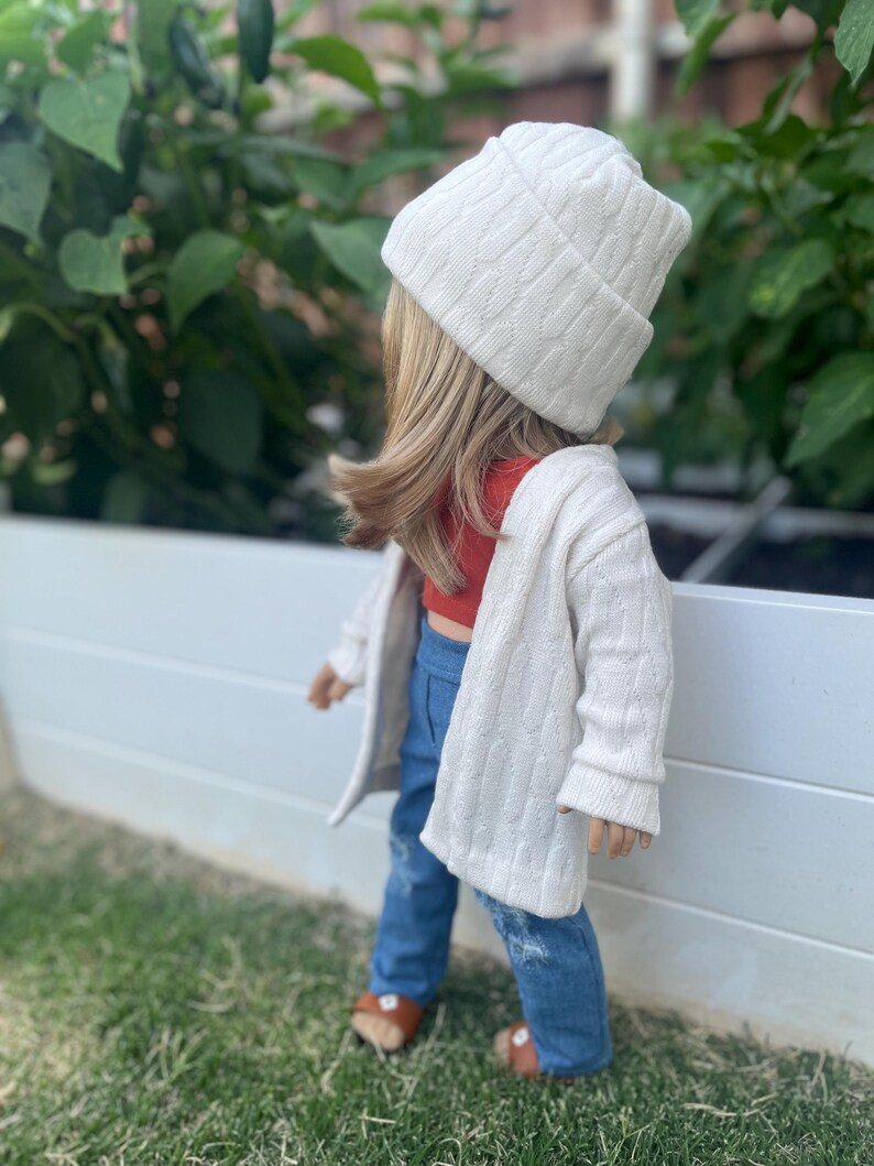 Op de afbeelding: Een pop die een witte kabelgebreide cardigan draagt, een rood topje, blauwe jeans en een witte kabelgebreide beanie.