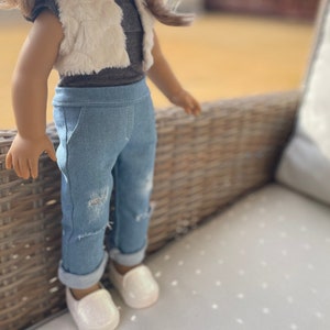 May include: A doll wearing a light brown faux fur vest over a gray shirt and blue denim jeans with distressed details. The doll is wearing white shoes.