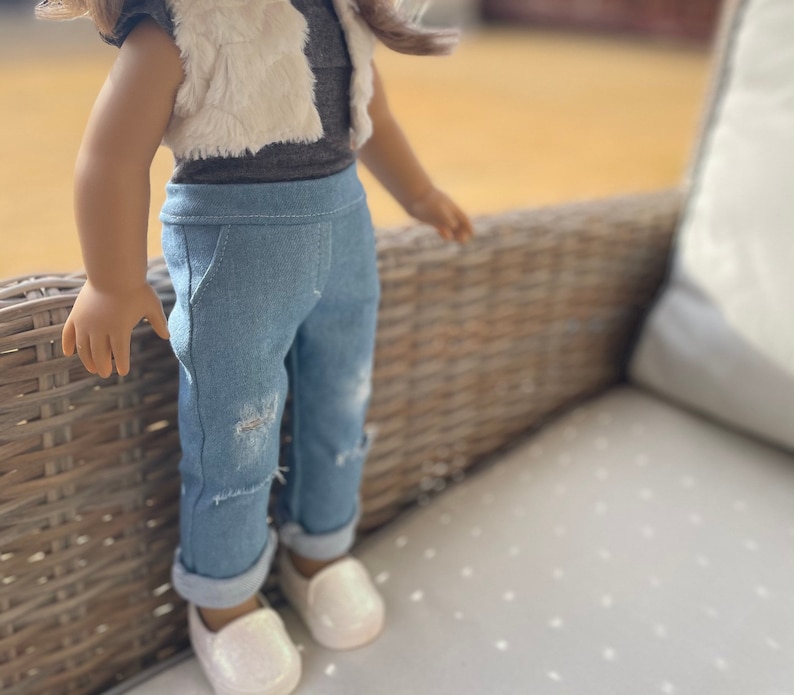 May include: A doll wearing a light brown faux fur vest over a gray shirt and blue denim jeans with distressed details. The doll is wearing white shoes.