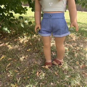 Peut inclure: Une poup&eacute;e v&ecirc;tue d'un haut court vert clair avec des d&eacute;tails de marguerites blanches et d'un short en jean bleu. Le short a une fermeture boutonn&eacute;e et des revers. La poup&eacute;e porte des sandales marron.