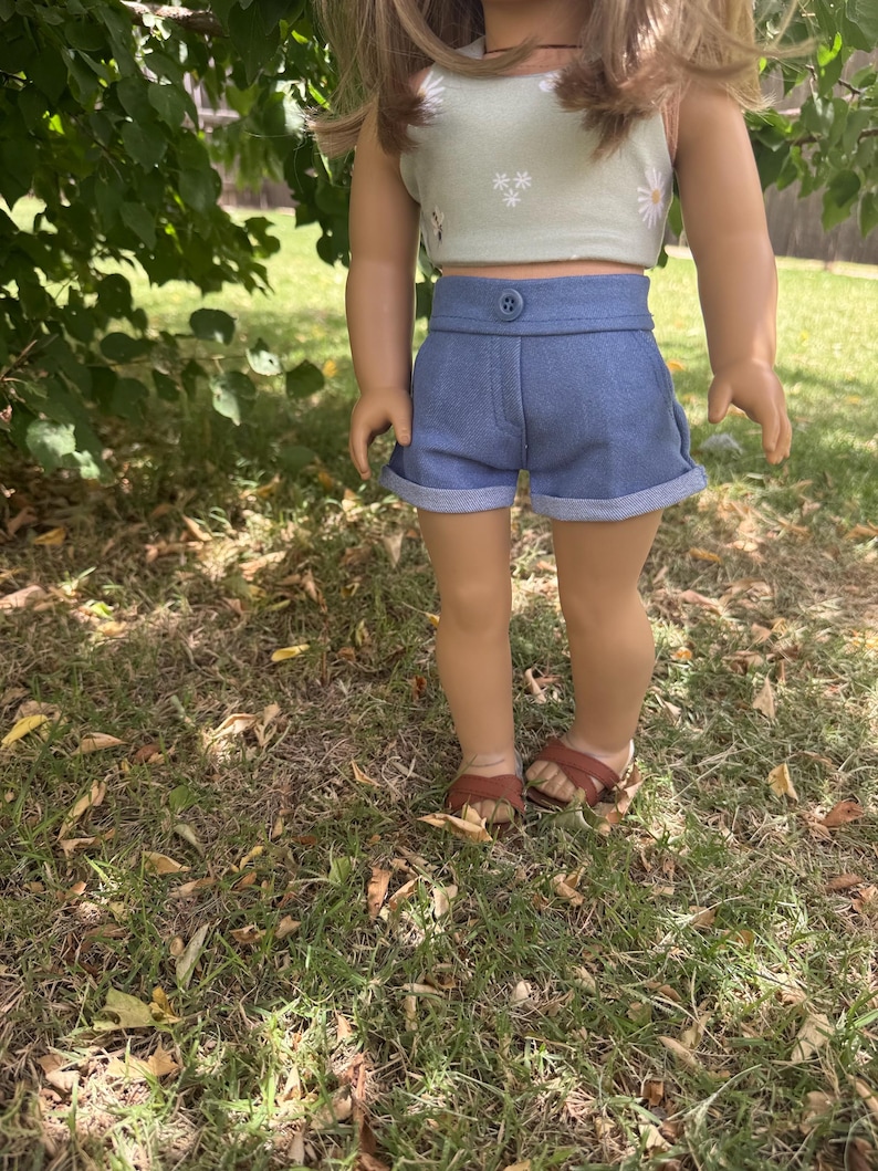 Peut inclure: Une poup&eacute;e v&ecirc;tue d'un haut court vert clair avec des d&eacute;tails de marguerites blanches et d'un short en jean bleu. Le short a une fermeture boutonn&eacute;e et des revers. La poup&eacute;e porte des sandales marron.