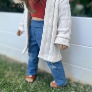 Op de afbeelding: Een pop die een witte cardigan, een rood kort topje, een blauwe spijkerbroek en bruine sandalen draagt. De pop staat voor een wit hek met groen gras op de achtergrond.