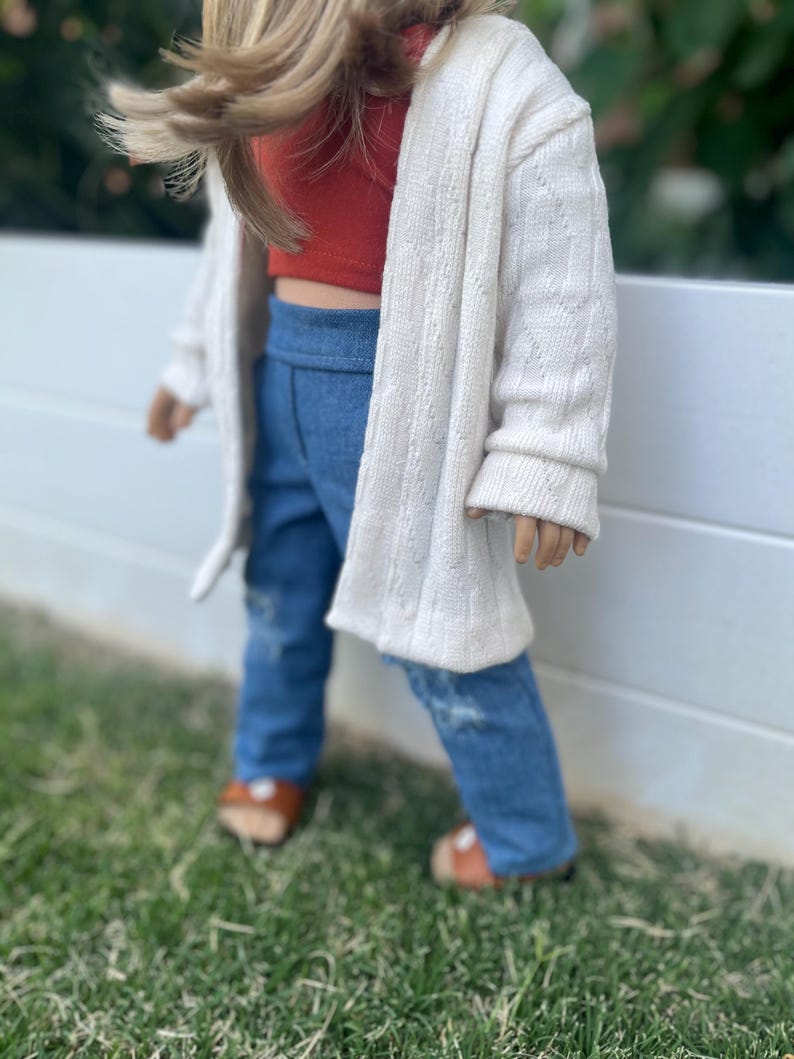 Op de afbeelding: Een pop die een witte cardigan, een rood kort topje, een blauwe spijkerbroek en bruine sandalen draagt. De pop staat voor een wit hek met groen gras op de achtergrond.