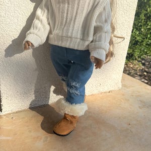 Puede incluir: Una mu&ntilde;eca con un su&eacute;ter blanco, jeans azules con roturas y botas de piel sint&eacute;tica marr&oacute;n.