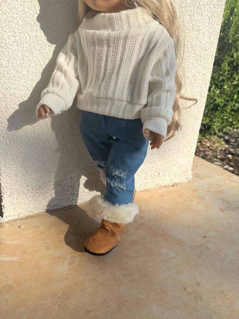 Puede incluir: Una mu&ntilde;eca con un su&eacute;ter blanco, jeans azules con roturas y botas de piel sint&eacute;tica marr&oacute;n.