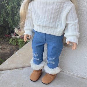 Puede incluir: Una mu&ntilde;eca con un su&eacute;ter blanco de cuello alto y jeans azules con detalles desgastados. La mu&ntilde;eca lleva botas marrones con ribete de piel blanca.