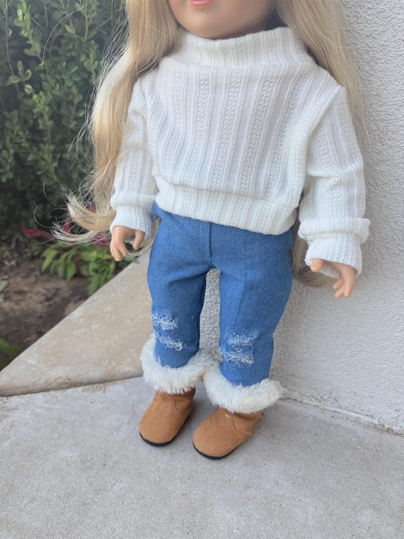 Puede incluir: Una mu&ntilde;eca con un su&eacute;ter blanco de cuello alto y jeans azules con detalles desgastados. La mu&ntilde;eca lleva botas marrones con ribete de piel blanca.
