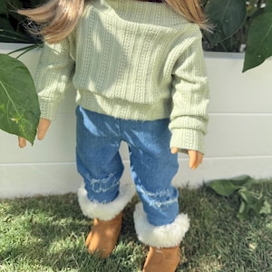 Puede incluir: Una mu&ntilde;eca con un su&eacute;ter verde claro y pantalones vaqueros azules con ribete de piel blanca en la parte inferior. La mu&ntilde;eca lleva botas marrones.
