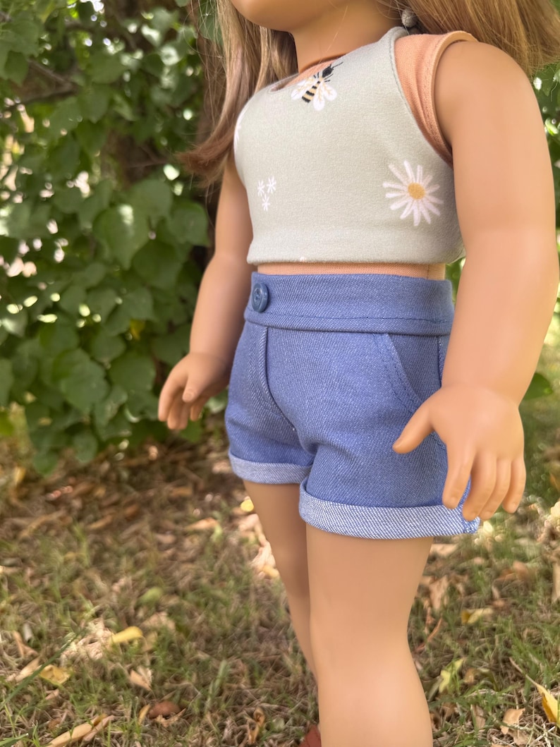 Peut inclure: Une poup&eacute;e v&ecirc;tue d'un haut court vert clair orn&eacute; d'un motif d'abeille et de marguerite, associ&eacute; &agrave; un short en jean bleu. Le short a un ourlet roul&eacute; et une fermeture boutonn&eacute;e. La tenue est con&ccedil;ue pour une poup&eacute;e.