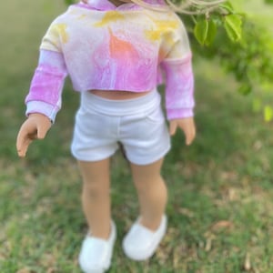 K&ouml;nnte beinhalten: Ein tie-dye-farbener, verk&uuml;rzter Hoodie mit langen &Auml;rmeln und wei&szlig;en Shorts. Der Hoodie hat ein gelbes, oranges und pinkfarbenes Farbschema. Die Shorts sind schlicht wei&szlig;.