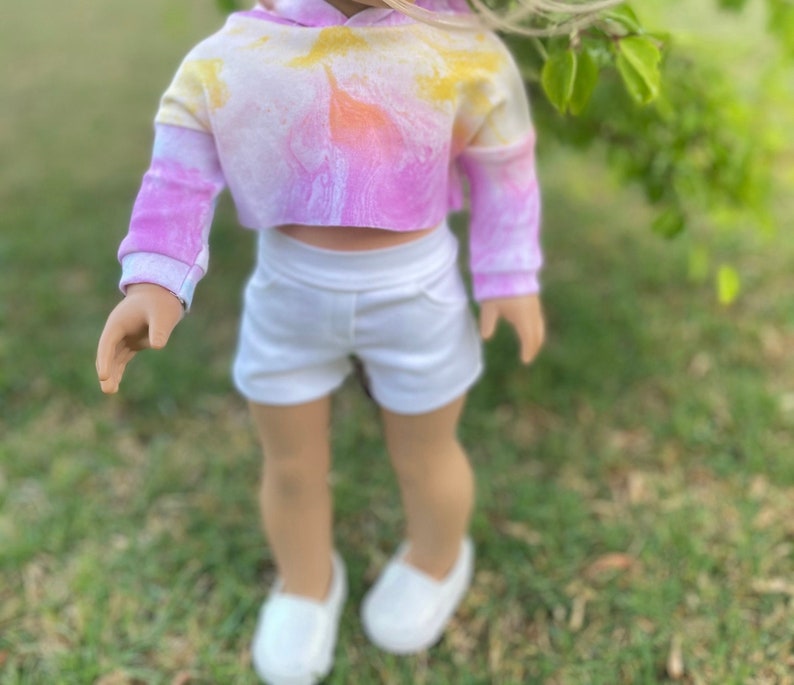 K&ouml;nnte beinhalten: Ein tie-dye-farbener, verk&uuml;rzter Hoodie mit langen &Auml;rmeln und wei&szlig;en Shorts. Der Hoodie hat ein gelbes, oranges und pinkfarbenes Farbschema. Die Shorts sind schlicht wei&szlig;.