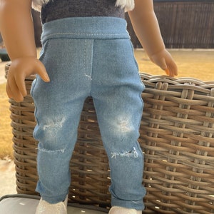 May include: A pair of light blue denim jeans with distressed details. The jeans have a straight leg fit and are worn by a doll. The doll is wearing a white fur vest and a gray shirt.