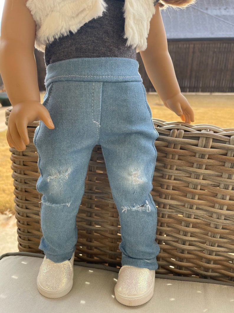 May include: A pair of light blue denim jeans with distressed details. The jeans have a straight leg fit and are worn by a doll. The doll is wearing a white fur vest and a gray shirt.