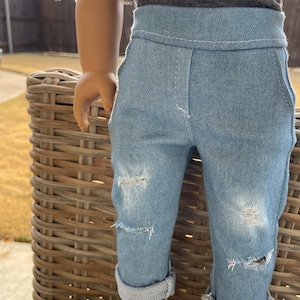 May include: A doll wearing a white ruffled shirt, a gray shirt, blue denim jeans with distressed details, and white sparkly shoes.