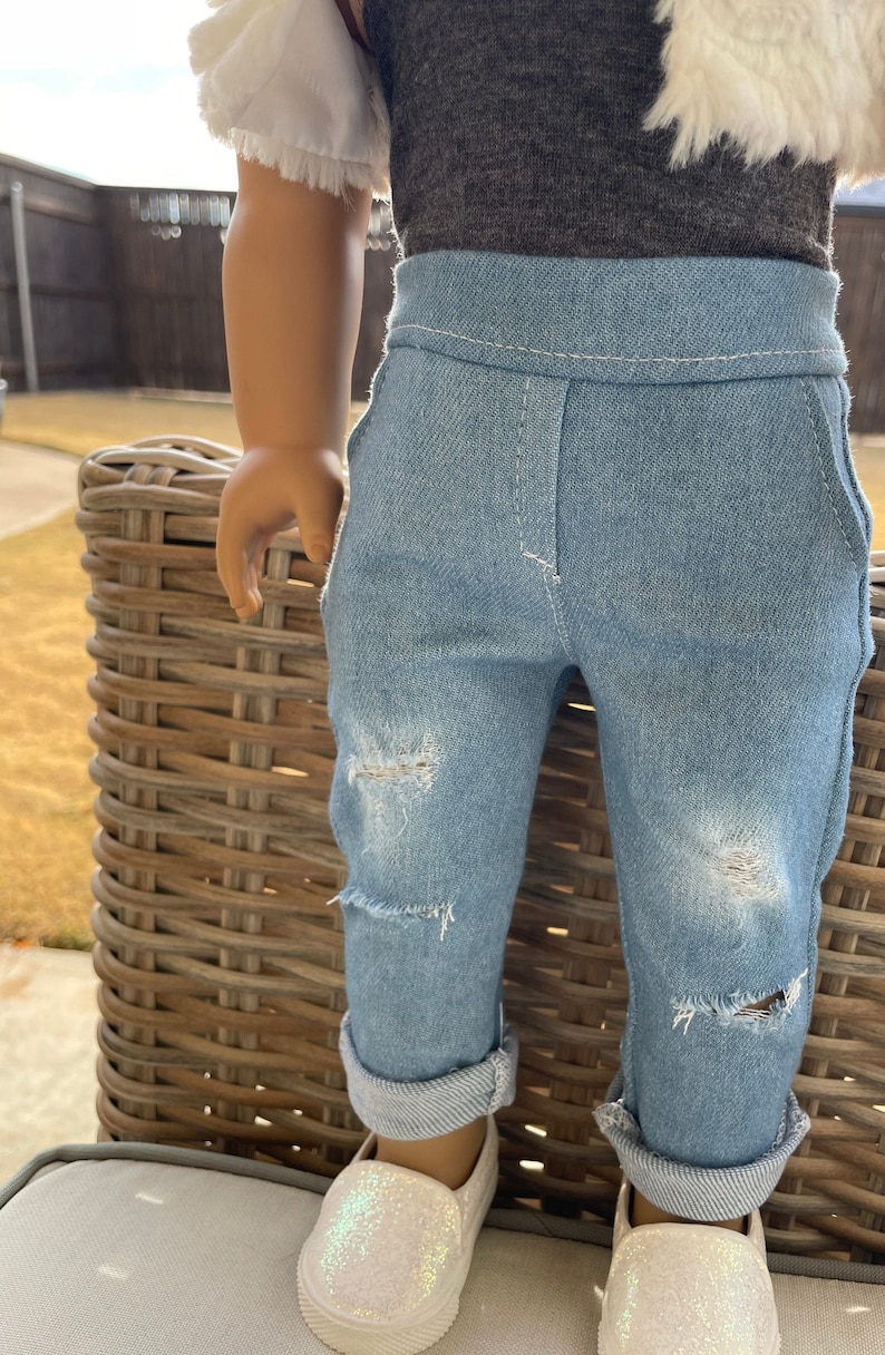 May include: A doll wearing a white ruffled shirt, a gray shirt, blue denim jeans with distressed details, and white sparkly shoes.