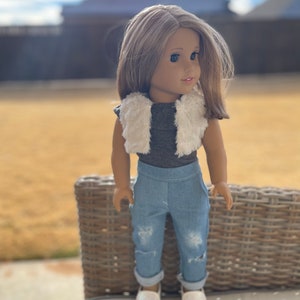 May include: A doll with brown hair and brown eyes wearing a white faux fur vest, a gray shirt, blue jeans with distressed details, and white shoes.