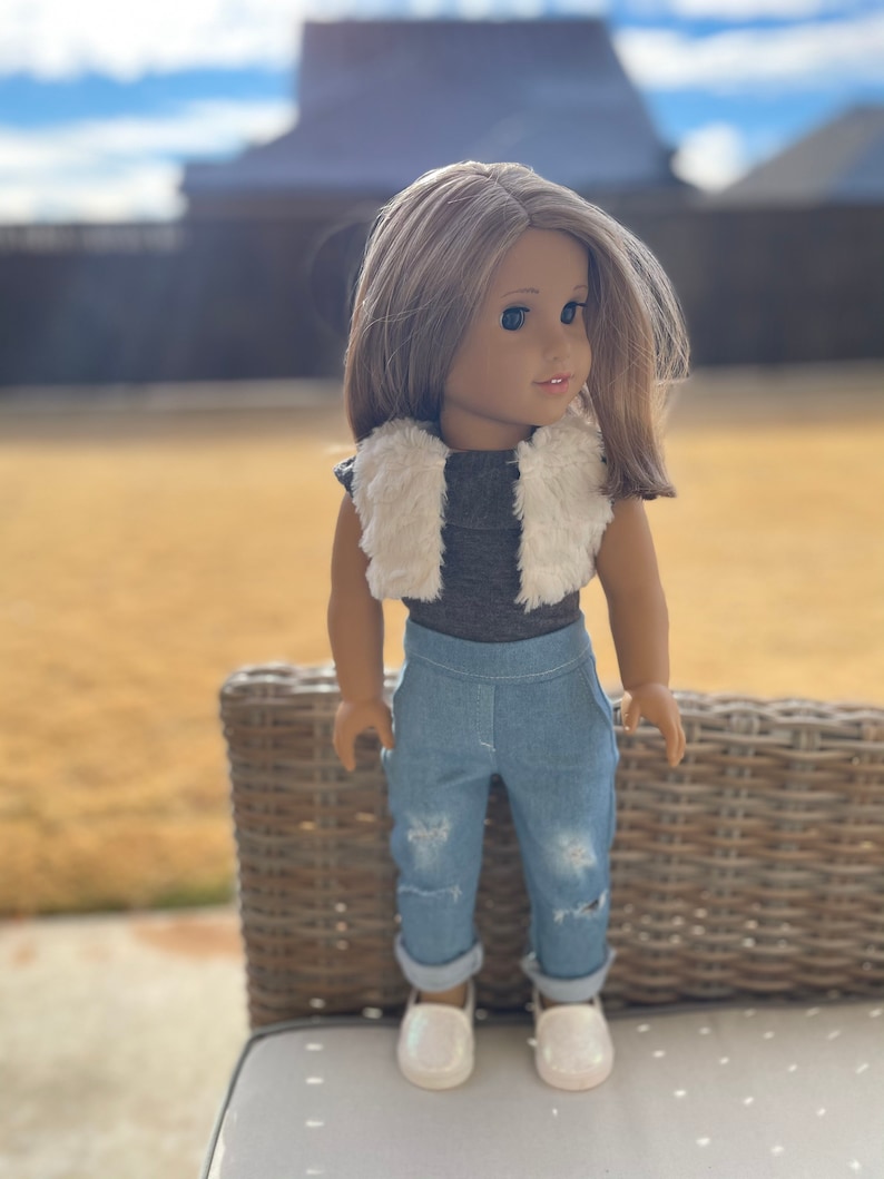 May include: A doll with brown hair and brown eyes wearing a white faux fur vest, a gray shirt, blue jeans with distressed details, and white shoes.
