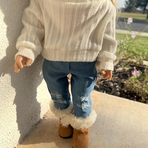 Puede incluir: Un su&eacute;ter de punto blanco con mangas largas y cuello alto, combinado con jeans azul denim y botas marrones de piel sint&eacute;tica.