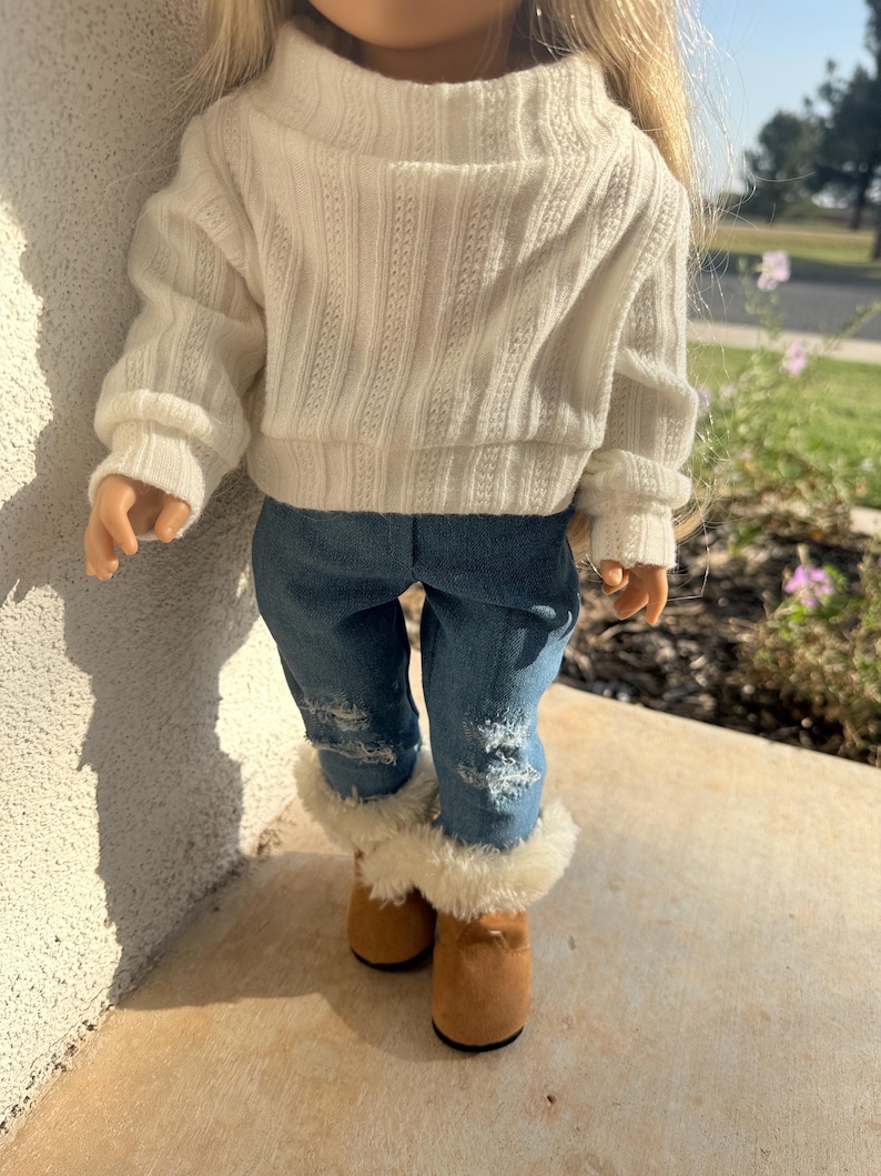 Puede incluir: Un su&eacute;ter de punto blanco con mangas largas y cuello alto, combinado con jeans azul denim y botas marrones de piel sint&eacute;tica.