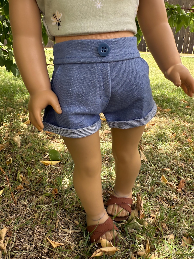 Peut inclure: Shorts en jean bleu avec fermeture boutonn&eacute;e et revers aux ourlets. Le short est associ&eacute; &agrave; un haut court vert clair avec un motif d'abeille. La poup&eacute;e porte des sandales marron. La tenue est con&ccedil;ue pour une poup&eacute;e.