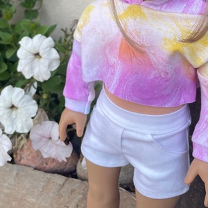 K&ouml;nnte beinhalten: Ein tie-dye-farbener, verk&uuml;rzter Hoodie mit langen &Auml;rmeln in den Farben Pink, Gelb und Orange. Der Hoodie wird mit einer wei&szlig;en Shorts kombiniert.