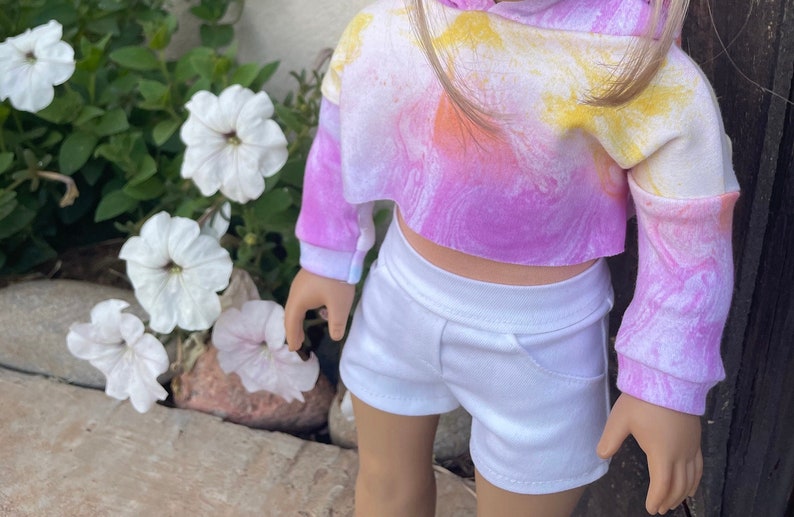 K&ouml;nnte beinhalten: Ein tie-dye-farbener, verk&uuml;rzter Hoodie mit langen &Auml;rmeln in den Farben Pink, Gelb und Orange. Der Hoodie wird mit einer wei&szlig;en Shorts kombiniert.
