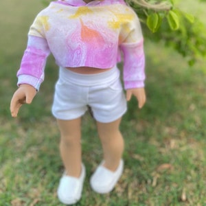 K&ouml;nnte beinhalten: Eine Puppe tr&auml;gt ein pinkfarbenes und gelbes tie-dye-farbenes, bauchfreies Hoodie und wei&szlig;e Shorts. Die Puppe steht auf einer grasbewachsenen Oberfl&auml;che.
