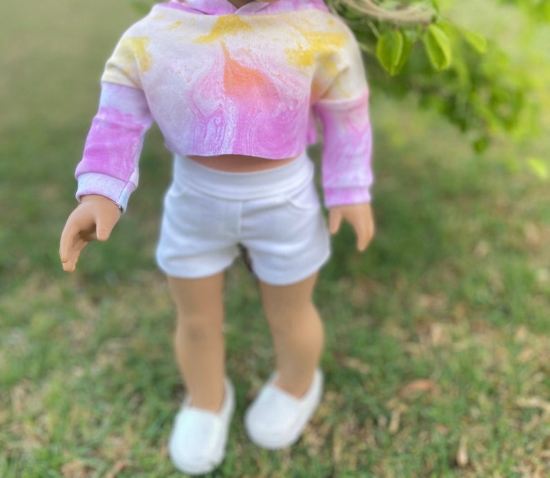 K&ouml;nnte beinhalten: Eine Puppe tr&auml;gt ein pinkfarbenes und gelbes tie-dye-farbenes, bauchfreies Hoodie und wei&szlig;e Shorts. Die Puppe steht auf einer grasbewachsenen Oberfl&auml;che.