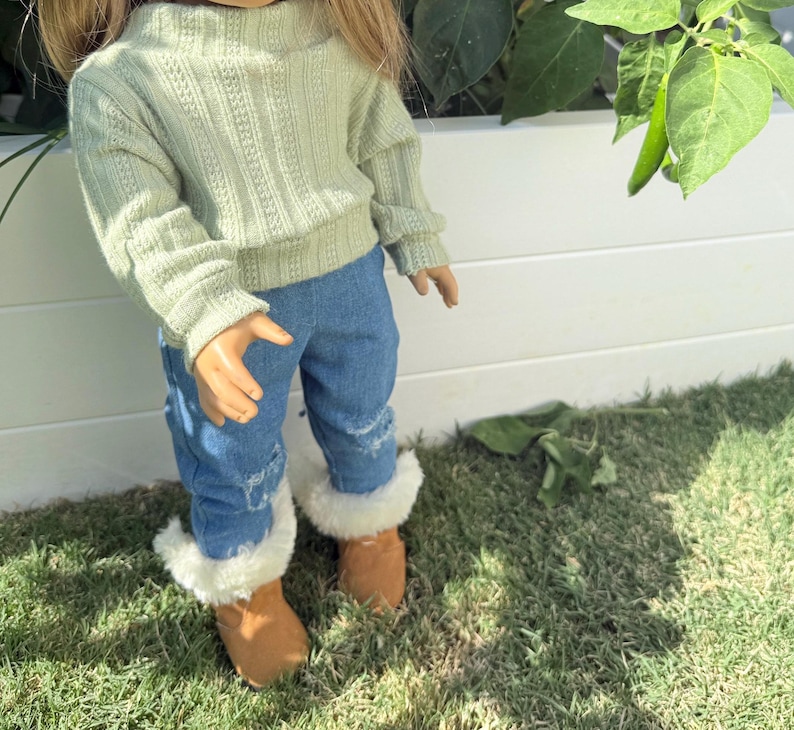 Puede incluir: Una mu&ntilde;eca con un su&eacute;ter verde claro y jeans azules con detalles desgastados. La mu&ntilde;eca lleva botas de piel sint&eacute;tica marr&oacute;n.
