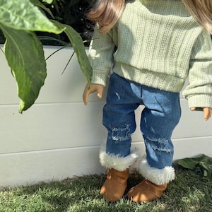 Puede incluir: Una mu&ntilde;eca con un su&eacute;ter verde claro y jeans azules con detalles desgastados. La mu&ntilde;eca lleva botas de piel sint&eacute;tica marr&oacute;n.